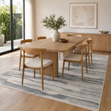 A modern dining room scene featuring a light oak wood rectangular table surrounded by six matching wooden chairs with cream upholstered seats, all arranged on a blue and gray abstract patterned area rug. The room has light wood flooring, a large window looking out onto greenery, and a minimalist sideboard in the background.