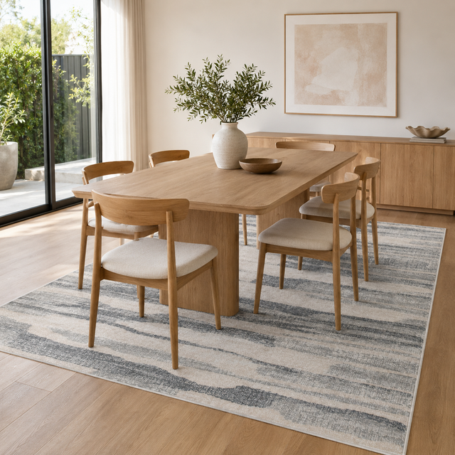 A modern dining room scene featuring a light oak wood rectangular table surrounded by six matching wooden chairs with cream upholstered seats, all arranged on a blue and gray abstract patterned area rug. The room has light wood flooring, a large window looking out onto greenery, and a minimalist sideboard in the background.